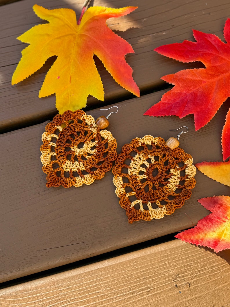 May include: A pair of handmade crochet earrings shaped like leaves, featuring a spiral pattern in shades of brown and cream yarn. Each earring is accented with a small, light brown wooden bead and attached to a silver-toned metal fishhook ear wire.