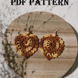 May include: A pair of heart-shaped crochet earrings featuring a variegated brown and cream yarn pattern with a small wooden bead at the top of each earring. The text PDF PATTERN is displayed above the earrings in a black serif font.