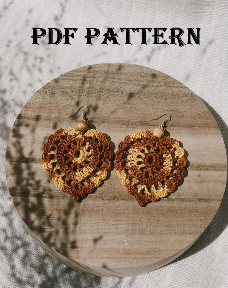 May include: A pair of heart-shaped crochet earrings featuring a variegated brown and cream yarn pattern with a small wooden bead at the top of each earring. The text PDF PATTERN is displayed above the earrings in a black serif font.