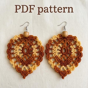 May include: A pair of heart-shaped crochet earrings featuring a brown and cream color scheme with a central wooden bead and silver-toned metal hooks. The text PDF pattern is displayed above the earrings in a brown sans-serif font.