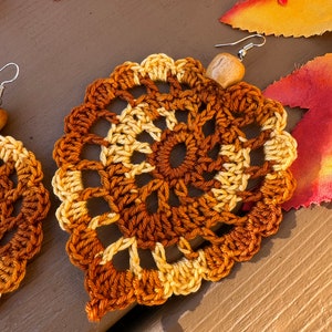 May include: A pair of crochet earrings shaped like leaves, featuring a variegated color pattern of dark brown, light brown, and cream yarn. Each earring is accented with a single light-colored wooden bead at the top and a silver-toned metal hook.