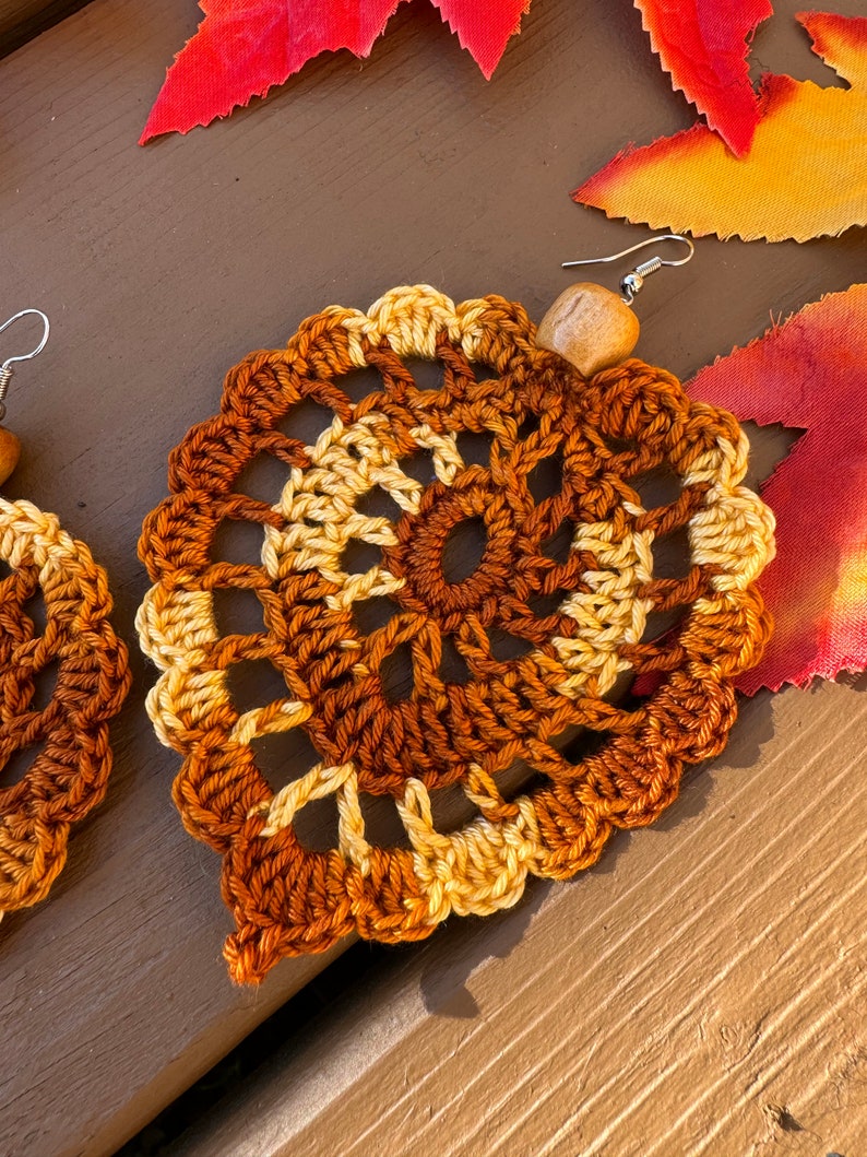 May include: A pair of crochet earrings shaped like leaves, featuring a variegated color pattern of dark brown, light brown, and cream yarn. Each earring is accented with a single light-colored wooden bead at the top and a silver-toned metal hook.