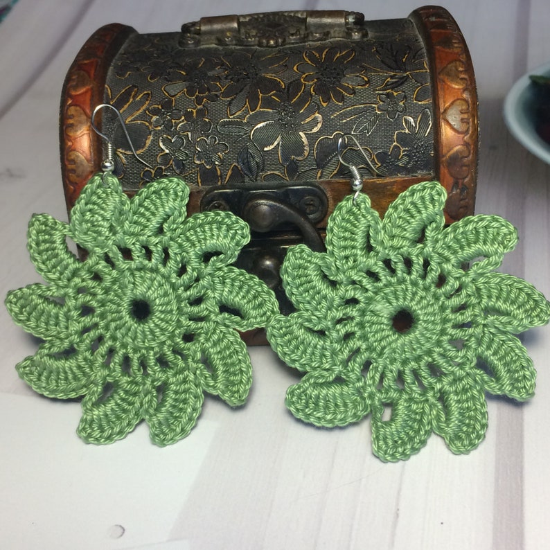 Pu&ograve; includere: Un paio di orecchini a forma di fiore di colore verde, realizzati a uncinetto, con ganci argentati. Gli orecchini sono delicati e intricati, con un design floreale.
