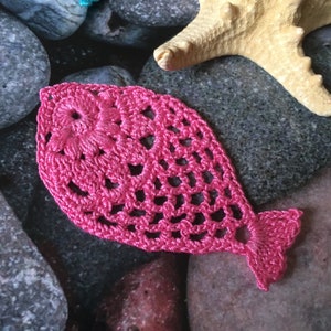 May include: A pink crocheted fish with a detailed design. The fish is made with a delicate, lacy stitch and has a rounded body and a tail fin.