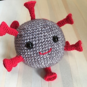May include: A gray crocheted plush toy shaped like a virus with red crocheted spikes and a smiling face. The toy has black eyes and a red stitched mouth.