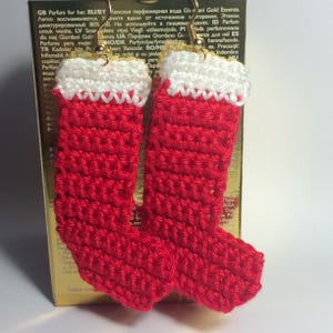 May include: A pair of handmade crochet earrings shaped like miniature Christmas stockings. Each stocking features a vibrant red body with a white cuff at the top, suspended from gold-toned metal earring hooks for pierced ears.