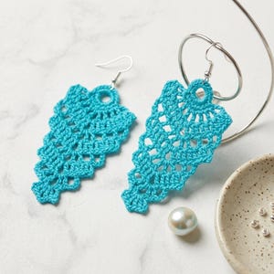 May include: A pair of bright turquoise crochet earrings featuring an intricate, openwork lace pattern in a triangular, leaf-like shape. Each earring is attached to a silver-toned metal fishhook wire, showcasing detailed thread work and craftsmanship.