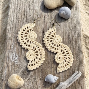 May include: A pair of cream-colored crochet earrings featuring an intricate, openwork fan-like pattern. Each earring is suspended from a simple bronze-toned metal hook, showcasing a delicate, lightweight design with a repeating circular motif.