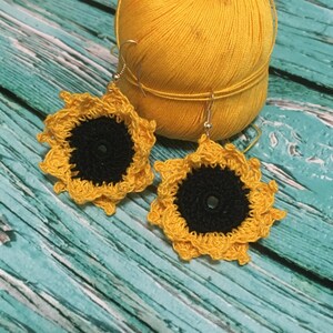 May include: A pair of handmade crochet earrings shaped like sunflowers, featuring a dark black circular center surrounded by bright yellow petals. Each earring is attached to a gold-toned metal hook for hanging, showcasing intricate yarn work.