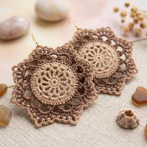 May include: A pair of round crochet earrings featuring a central light beige starburst pattern surrounded by concentric rings of dark brown thread. The earrings have a delicate, openwork lace design with scalloped edges and gold-toned metal hooks.