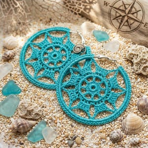 May include: A pair of round hoop earrings featuring intricate teal crochet work in a starburst pattern. Each earring is attached to a silver-toned metal hoop and includes a small, metallic ammonite fossil charm dangling from the top edge.