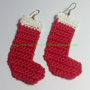 May include: A pair of handmade crochet earrings shaped like Christmas stockings. Each earring features a vibrant red body with a white cuff at the top and a gold-toned metal fishhook ear wire. The text www.etsy.com/shop/CreativeAccessorise is visible.
