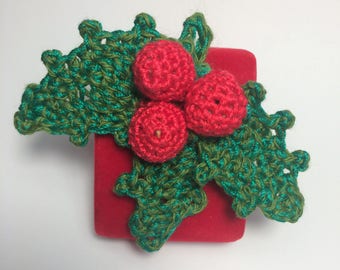 May include: A crocheted holly sprig featuring three round red berries and two textured green leaves with jagged edges, resting on top of a small, square, red velvet box. The yarn work displays visible loops and a consistent stitch pattern.