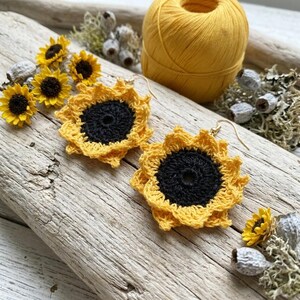 May include: A pair of handmade crochet sunflower earrings featuring bright yellow petals and a dark brown center. Each earring is crafted with fine yarn, showcasing intricate stitching and a gold-toned metal hook for attachment to the ear.