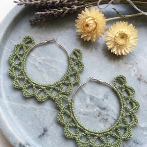 May include: A pair of sage green crochet hoop earrings featuring a delicate scalloped edge pattern. The earrings are constructed with silver-toned metal hoops and intricate green thread work that forms a decorative, repeating lace-like border.