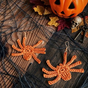 May include: Two orange crochet spider earrings with gold hooks. The earrings are set on a wooden surface with Halloween decorations, including a pumpkin, skull, and a faux spider web.