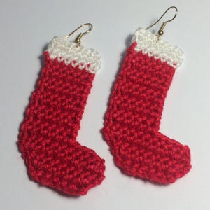 May include: A pair of handmade crochet earrings shaped like Christmas stockings. Each earring features a vibrant red body with a white cuff at the top and a gold-toned metal fishhook ear wire for attachment, showcasing a textured yarn construction.