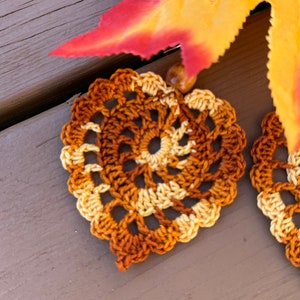 May include: A pair of heart-shaped crochet earrings featuring a variegated yarn pattern in shades of brown, tan, and cream. Each earring has a scalloped edge and is accented with a small, round, light brown bead at the top attachment point.
