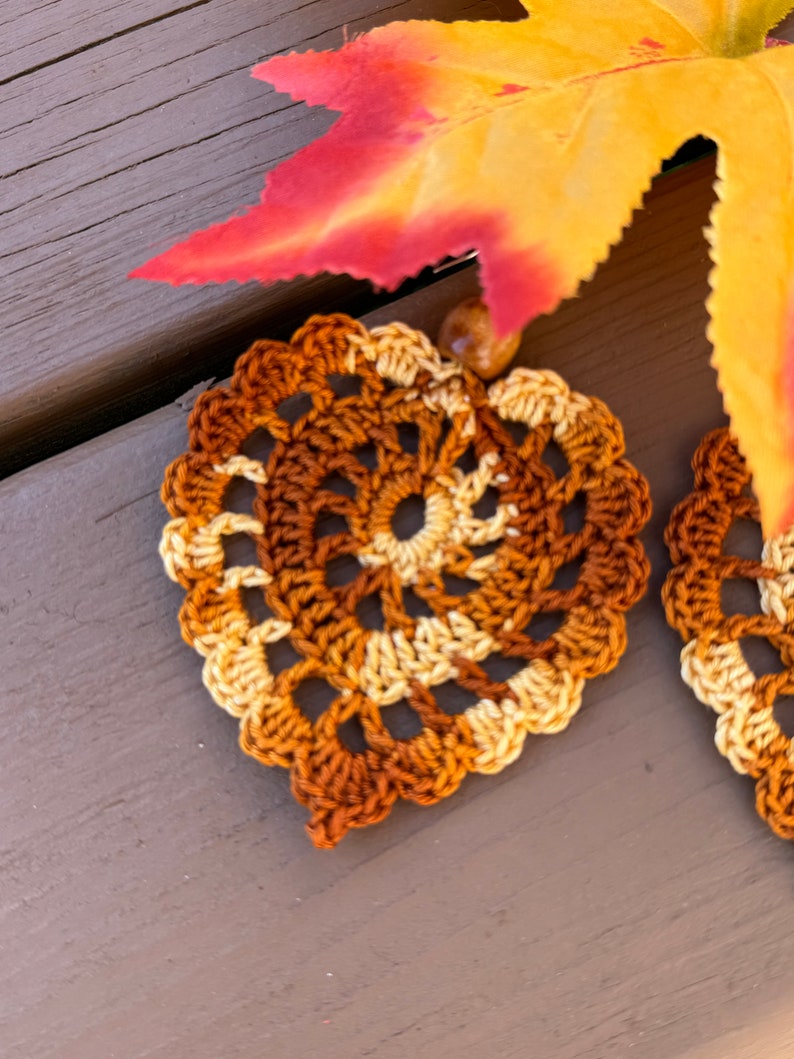 May include: A pair of heart-shaped crochet earrings featuring a variegated yarn pattern in shades of brown, tan, and cream. Each earring has a scalloped edge and is accented with a small, round, light brown bead at the top attachment point.
