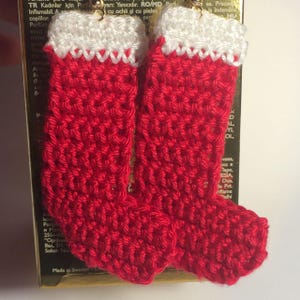 May include: A pair of handmade crochet earrings shaped like miniature Christmas stockings. Each stocking features a vibrant red body with a textured white cuff at the top. The earrings are suspended from gold-toned metal hooks for attachment.