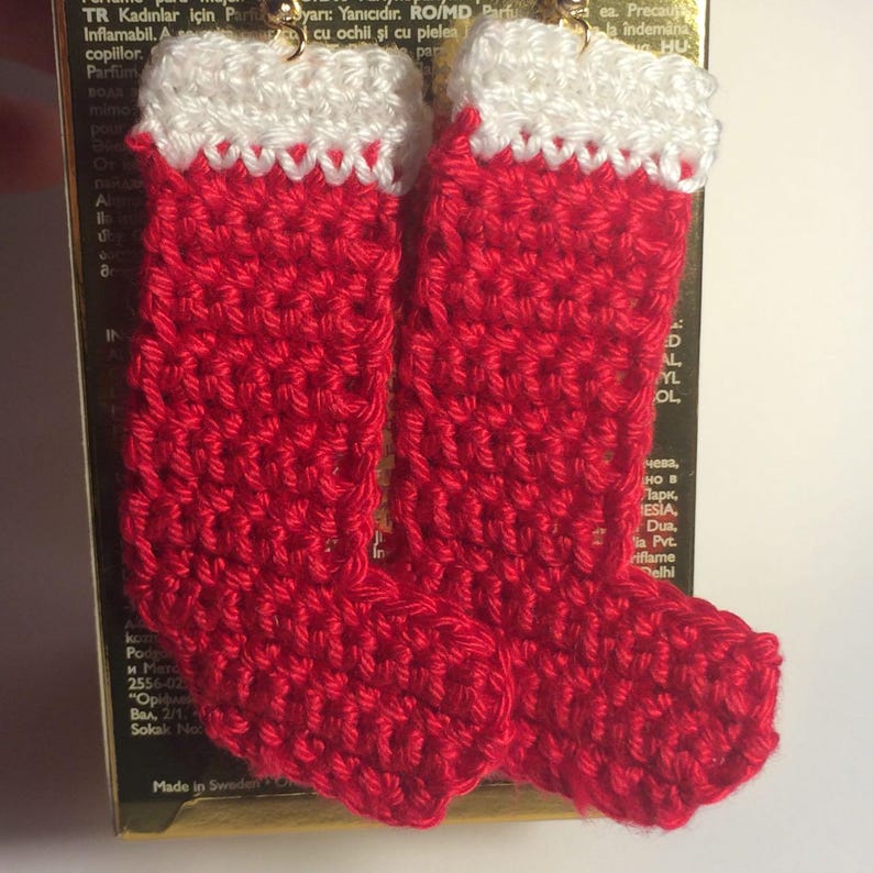 May include: A pair of handmade crochet earrings shaped like miniature Christmas stockings. Each stocking features a vibrant red body with a textured white cuff at the top. The earrings are suspended from gold-toned metal hooks for attachment.