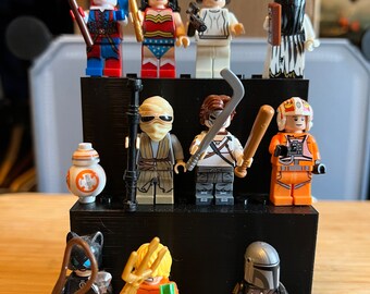 May include: A collection of 11 minifigures displayed on a black tiered stand. The minifigures include characters from Star Wars, DC Comics, and Marvel Comics. The top tier features Harley Quinn, Wonder Woman, Princess Leia, and a black-haired female character. The middle tier features a character with a brown beard and a black mask, a character with a black mask and a brown beard, a character with a white mask and a brown beard, and a character with a white helmet and a brown beard. The bottom tier features Catwoman, Aquaman, The Mandalorian, and Baby Yoda.