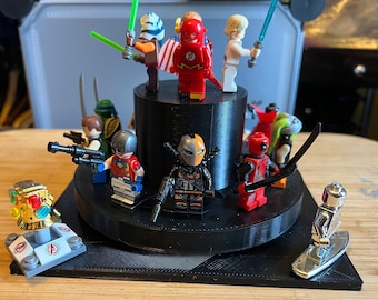 May include: A collection of superhero and villain minifigures, including Flash, Deadpool, and Deathstroke, displayed on a black tiered stand. The figures are made of plastic and have a variety of colors, including red, black, green, and yellow.