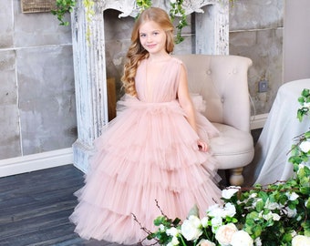 May include: A young girl wearing a pink tulle dress sits on a white chair in front of a fireplace decorated with white and green flowers. The floor is made of dark wood planks.