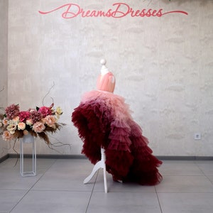 Könnte beinhalten: Ein rosa-burgunderfarbenes Ombre-Tüllkleid mit einem High-Low-Saum, das auf einer weißen Kleiderpuppe präsentiert wird. Das Kleid hat mehrere Tüllschichten und ist mit einer rosa Schleife in der Taille verziert. Das Kleid wird in einem weißen Raum mit einem Blumenarrangement und dem Text "Dreams Dresses" an der Wand ausgestellt.