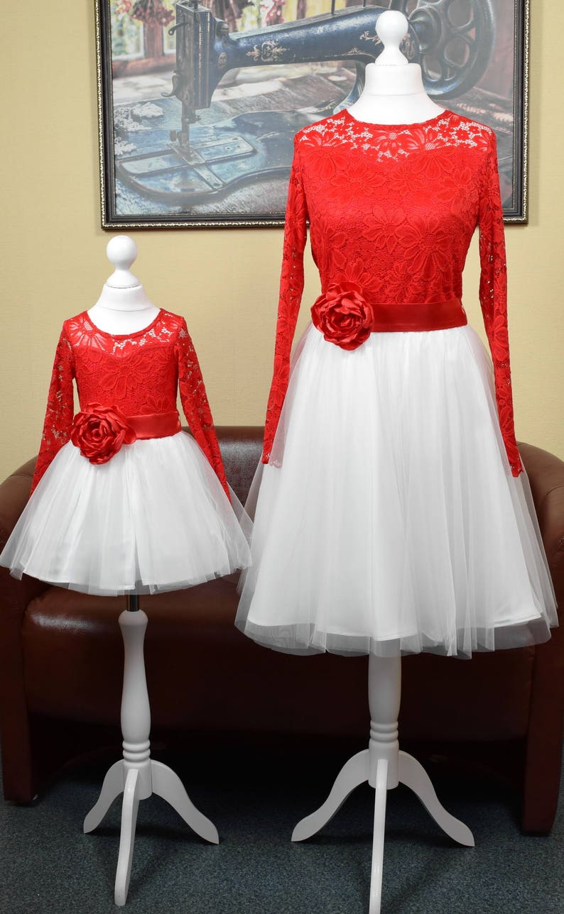 May include: Two red lace dresses with white tulle skirts and red satin sashes with a large red flower accent. The dresses are on white mannequins with white stands.