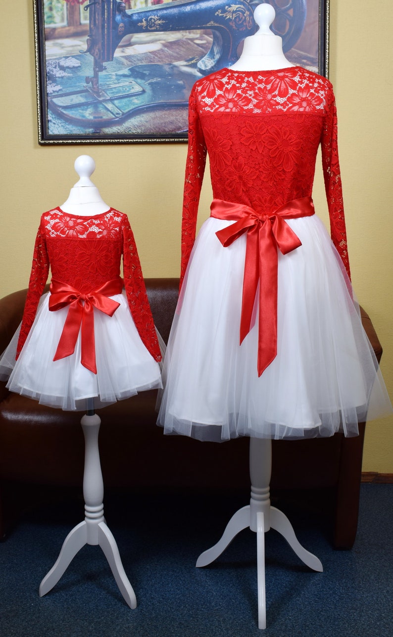 May include: Two red lace dresses with white tulle skirts and red satin bows. The dresses are on white mannequins with wooden stands.