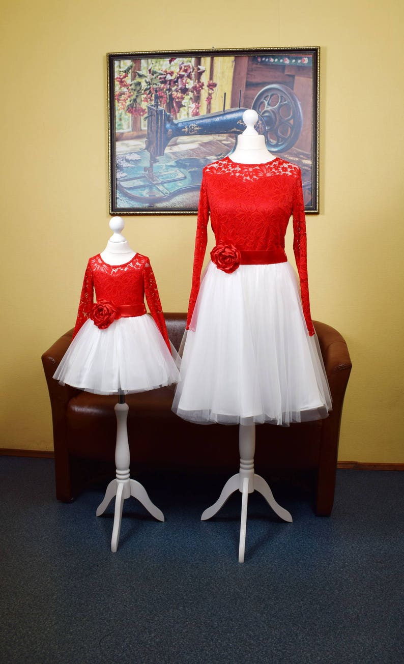 May include: Two red lace dresses with white tulle skirts and red flower accents. The dresses are on mannequins with white stands. The dresses are on a dark blue carpet in front of a brown leather couch.