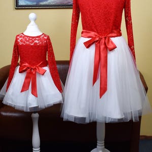 May include: Two red lace dresses with white tulle skirts and red satin bows. The dresses are on white mannequins with white stands.