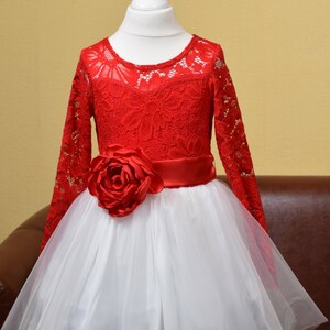 May include: A red lace dress with long sleeves and a white tulle skirt. The dress has a red satin ribbon around the waist and a large red rose on the front.