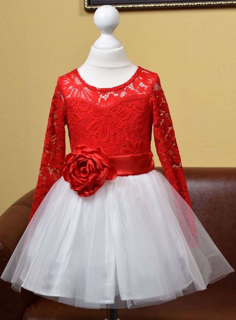 May include: A red lace dress with long sleeves and a white tulle skirt. The dress has a red satin ribbon around the waist and a large red rose on the front.