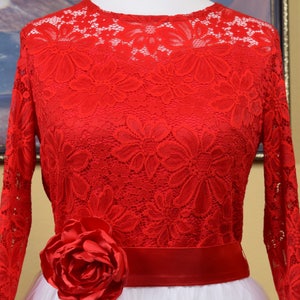 May include: Red lace dress with long sleeves and a white tulle skirt. The dress has a red satin sash and a large red rose on the front.