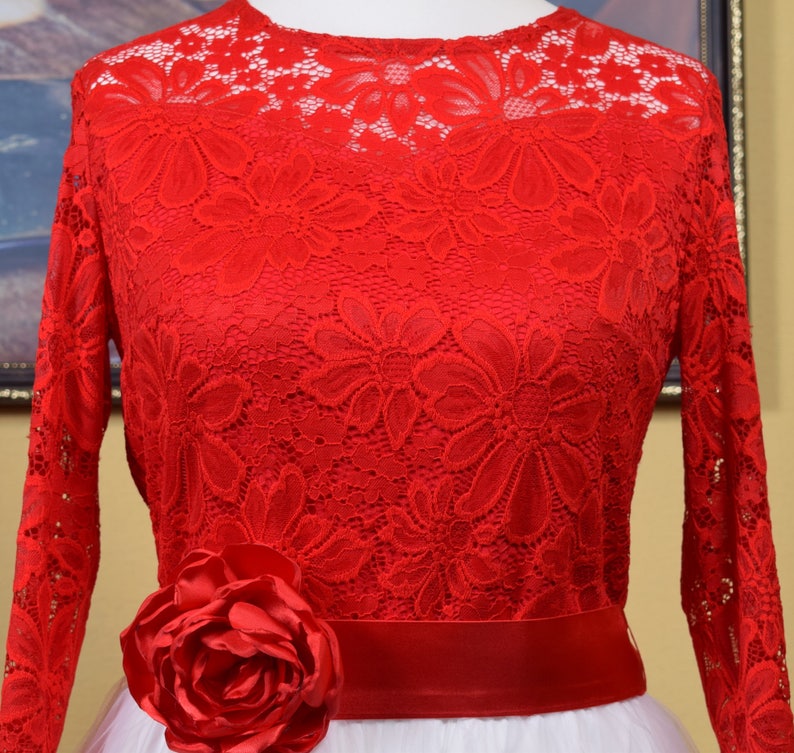 May include: Red lace dress with long sleeves and a white tulle skirt. The dress has a red satin sash and a large red rose on the front.