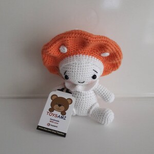 Op de afbeelding: Een gehaakt pluche speelgoed in de vorm van een paddenstoel met een wit lichaam en een oranje hoed. Het speelgoed zit op een wit oppervlak met een label waarop staat "TOYSAMi Handgemaakt met liefde @toysami www.etsy.com/shop/Toysami"