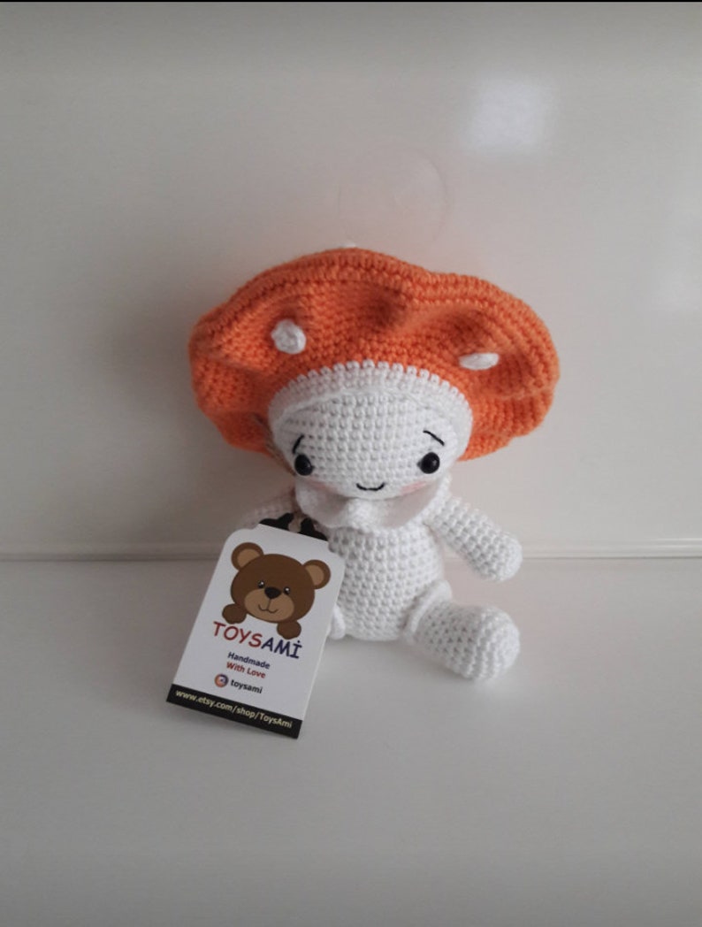 Op de afbeelding: Een gehaakt pluche speelgoed in de vorm van een paddenstoel met een wit lichaam en een oranje hoed. Het speelgoed zit op een wit oppervlak met een label waarop staat "TOYSAMi Handgemaakt met liefde @toysami www.etsy.com/shop/Toysami"