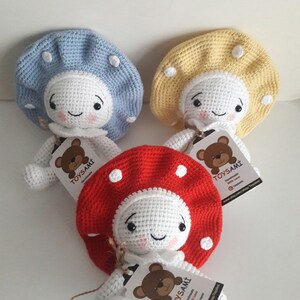 Op de afbeelding: Drie gehaakte paddenstoelpoppen met witte lichamen, glimlachende gezichten en verschillende gekleurde hoeden. De hoeden zijn blauw, geel en rood met witte stippen. Elke pop heeft een label met de tekst "TOYSAMİ Handgemaakt met liefde www.etsy.com/shop/toysami"