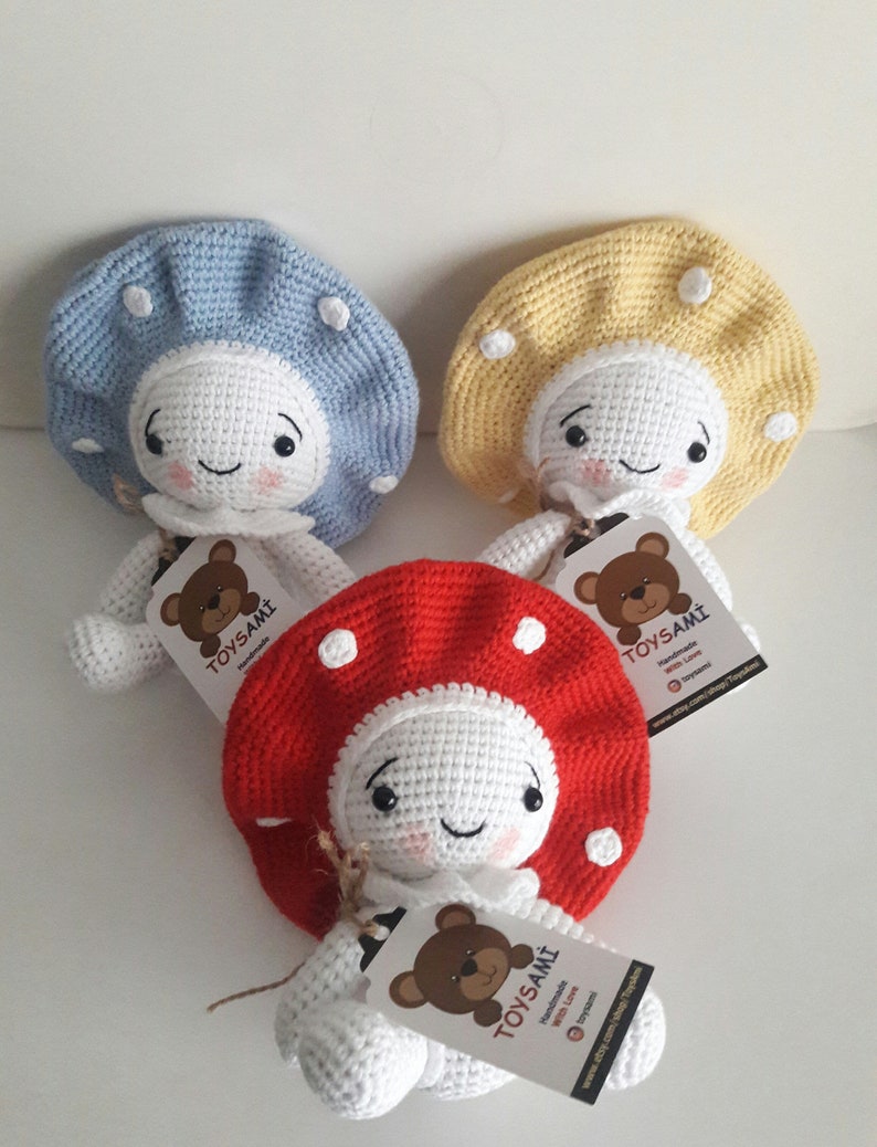 Op de afbeelding: Drie gehaakte paddenstoelpoppen met witte lichamen, glimlachende gezichten en verschillende gekleurde hoeden. De hoeden zijn blauw, geel en rood met witte stippen. Elke pop heeft een label met de tekst "TOYSAMİ Handgemaakt met liefde www.etsy.com/shop/toysami"