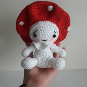 Op de afbeelding: Een gehaakt knuffeldiertje in de vorm van een paddenstoel met een rode dop en een wit lichaam. De paddenstoel heeft een glimlachend gezicht met zwarte ogen en roze wangen.