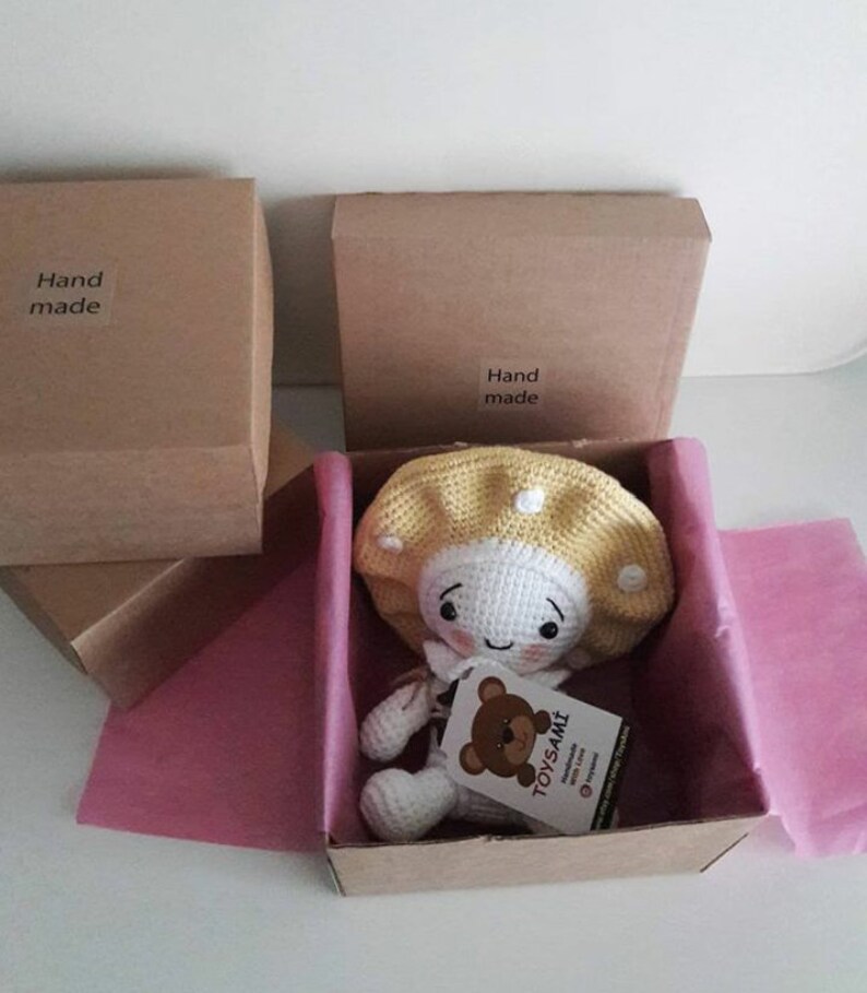 Op de afbeelding: Een gehaakte paddenstoelpop met een gele hoed en een wit lichaam. De pop zit in een bruine kartonnen doos met roze tissuepapier. De doos heeft een label met de tekst "Handgemaakt".