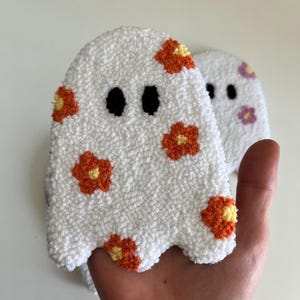 May include: A white ghost with a fuzzy texture and orange and yellow flowers embroidered on it. The ghost has black eyes and a simple, friendly expression.