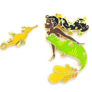 May include: Two enamel pins: a yellow seahorse and a mermaid. The seahorse is yellow with green and gold accents. The mermaid has brown skin, black hair with green accents, and a green dress and tail with yellow and orange details.