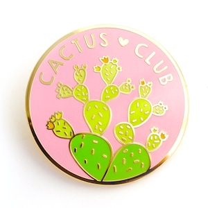 May include: A pink and gold enamel pin with the text "Cactus Club" and a cartoon image of a green cactus with yellow flowers.