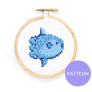 May include: A blue and white cross-stitch pattern of a cartoon fish in a wooden embroidery hoop. The hoop has a brass screw and knob. The text "PATTERN" is on a purple circle below the hoop.