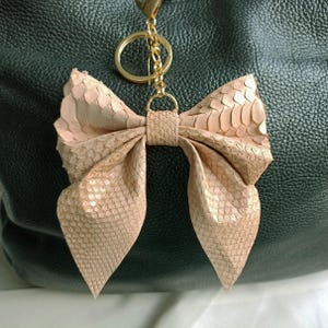 Könnte beinhalten: Ein hellrosa schlüsselanhänger in Schleifenform aus strukturiertem Material, möglicherweise Leder oder einem ähnlichen Material. Der Schlüsselanhänger hat einen goldfarbenen Ring und Verschluss. Der Hintergrund ist eine dunkelgrüne Ledertasche.