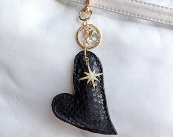May include: A black heart-shaped keychain with a gold starburst charm and a gold-colored clasp. The keychain is attached to a white leather bag. The heart has a textured, snake-like pattern.