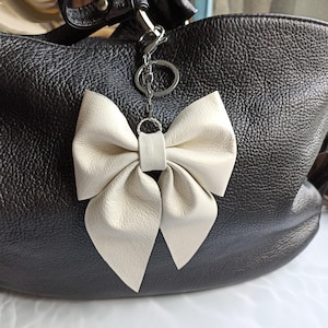 Könnte beinhalten: Eine schwarze Lederhandtasche mit einem cremefarbenen Leder-Schlüsselanhänger in Schleifenform. Die Schleife ist mit mehreren Falten versehen und hängt an einem silberfarbenen Metallring und -verschluss. Die Tasche hat eine strukturierte Oberfläche.